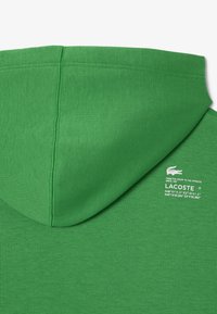 Felpa verde con una texture morbida, dotata di tasca a marsupio e patch con logo. Il design include un cappuccio con lacci e schiena senza cuciture.