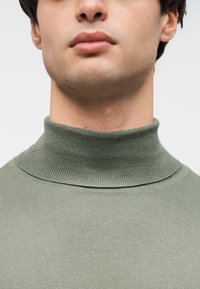 Gros plan sur le bas du visage et le cou d'une personne portant un col roulé vert côtelé, sur un fond clair uni.