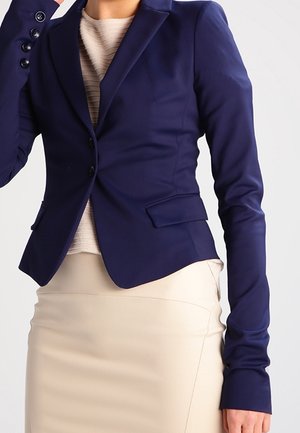 Vrouw draagt een aansluitend marineblauw blazer over een beige gestructureerde top en een lichtbeige kokerrok, staand tegen een effen lichte achtergrond.