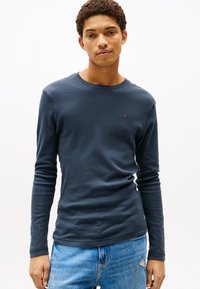 Granatowa koszulka z długim rękawem wykonana z miękkiej bawełny, z okrągłym dekoltem i małym logo na piersi, stylizowana z jasnoniebieskimi dżinsami.