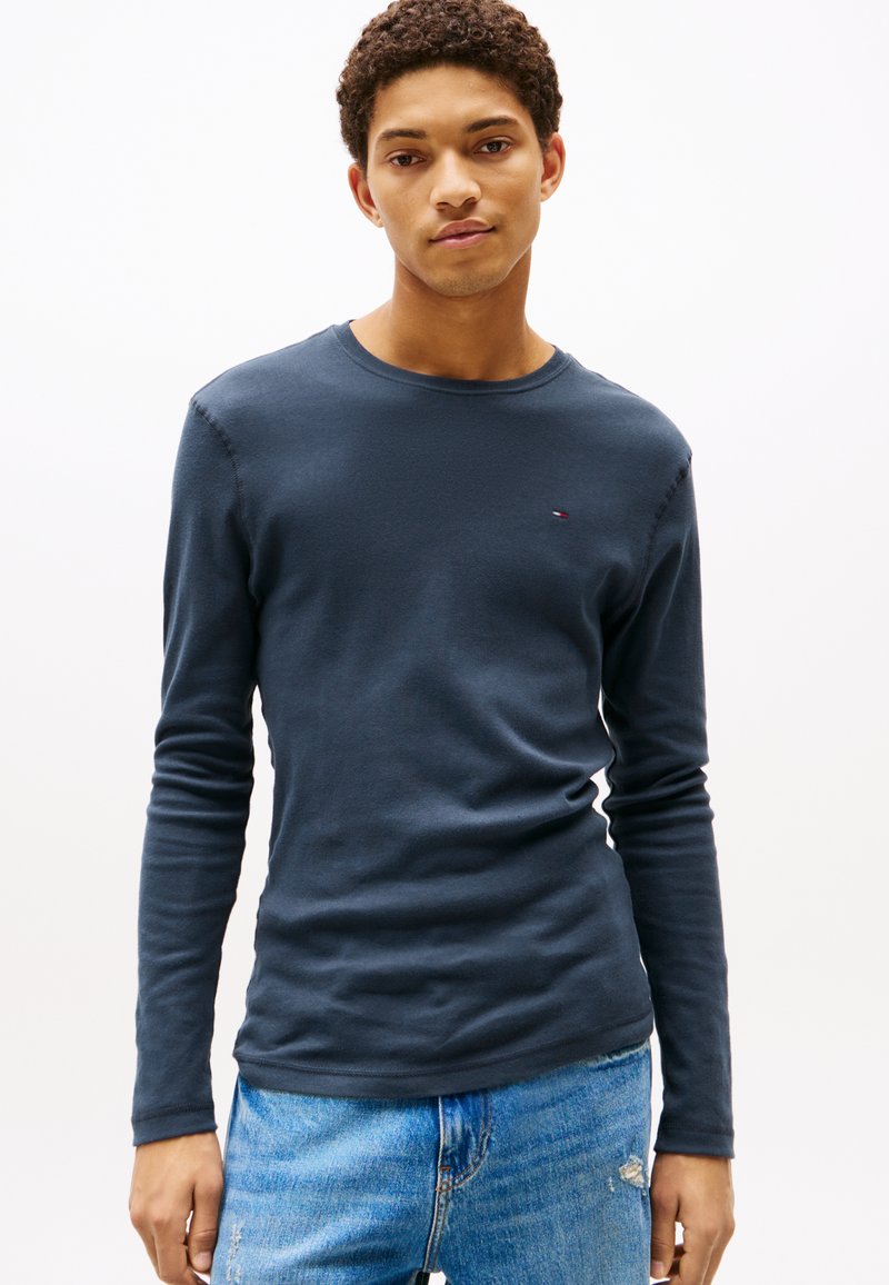 Granatowa koszulka z długim rękawem wykonana z miękkiej bawełny, z okrągłym dekoltem i małym logo na piersi, stylizowana z jasnoniebieskimi dżinsami.