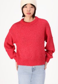 Rdeča pletena jopica s krožnim izrezom, spuščeni rameni in teksturiranimi kroglicami; v kombinaciji s svetlim jeans krilom in smetanasto kapo.