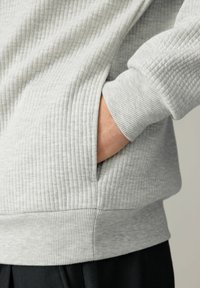 Detalle de sudadera gris de canalé que muestra la tela texturizada, un bolsillo lateral y el puño. El diseño presenta un patrón de rayas sutil y un dobladillo ajustado.