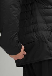 Juodas izoliuotas paltas su pūkuotu dizainu, turintis užtrauktuku užsegamą šoninę kišenę ir lygią tekstūrą. Rankovė turi prigludusį manžetą šilumai.