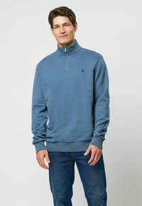 Kék, negyedcipzáras pulóver, puha anyagból, bordás szegéllyel és mandzsettával. Kis logóval a mellkason és hosszú ujjakkal rendelkezik. Sötét farmernadrágot visel.