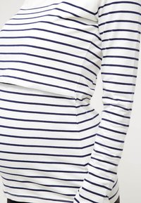 Personne enceinte portant une chemise blanche à manches longues avec des rayures horizontales bleu marine, vue de profil.
