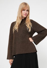 Suéter castanho tricotado com gola alta e mangas largas em canelado, apresentando um ajuste solto e uma textura suave, combinado com uma saia preta.