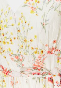 Blommig klänning i lätt tyg. Har en V-ringning, dämpade färger och ett bälte vid midjan med mönster av rosa och gula blommor.