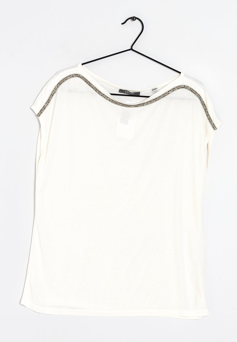 Bílý krátký rukáv tričko s volným střihem, s metalickým okrajem kolem krku a hladkým, lehkým materiálem. Zavěšeno na černém háčku.