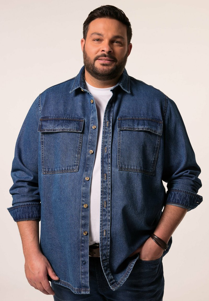 Camisa de mezclilla abotonada de color azul oscuro con dos bolsillos frontales, un cuello clásico y puños enrollados sobre una camiseta blanca. Llevada con jeans oscuros.