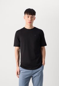 Černé tričko s krátkým rukávem vyrobené z měkké tkaniny, s minimalistickým designem a malým zeleným logem na hrudi.