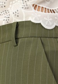 Olivengrønne bukser med hvide striber, der har flat front, sidelommer og en glat stofstruktur. Øverdelen viser broderet blonder.