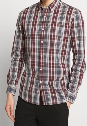 Homme portant une chemise à manches longues avec un motif à carreaux rouge, gris et blanc, et un pantalon noir, une main dans la poche.