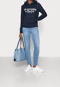 Sudadera con capucha navy y logo blanco, jeans skinny azules claros, zapatillas blancas y una bolsa tote azul claro con acentos azul oscuro.