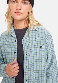 Groen en blauw geruit flanellen shirt met zwarte knopen, voorzien van een kraag en een borstzak, gelaagd over een donker T-shirt.