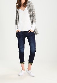 Grau-weiß gemusterter Cardigan über einem weißen V-Ausschnitt-Oberteil mit kreisförmigen Details, kombiniert mit dunklem Denim-Jeans und weißen Sneakers mit gelben Akzenten.