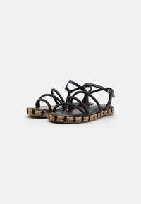 Sandales gladiateur noires avec des lanières tressées, semelle plate et garniture en raffia tissé, dotées d'une fermeture à boucle pour un maintien sécurisé.