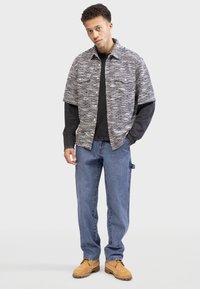 Chemise à manches courtes à motifs gris et noir, portant une couche noire unie en dessous, assortie à un jean bleu et des bottes beiges.