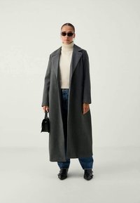 Ilgas pilkas vilnonis paltas su išpjauta apykakle, dėvimas virš kreminės spalvos raukšlėtos turtlenkės ir mėlynų plačių džinsų. Aksesuarai apima juodus saulės akinius ir nedidelę rankinę.
