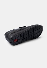 Chaussure en cuir noir avec une semelle en caoutchouc texturé arborant divers motifs de semelle et un accent circulaire rouge près du talon. Fermeture à strap Velcro.
