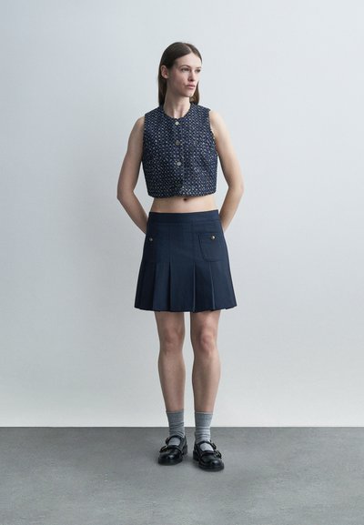 Γυναίκα που στέκεται με τα χέρια πίσω από την πλάτη, φορώντας ένα σκούρο μπλε χωρίς μανίκια κοντό κουμπωτό τοπ, φούστα με πιέτες και τσέπες, γκρι κάλτσες και μαύρα παπούτσια.