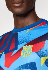 Kék sportmez, élénk grafikus mintázattal piros és sárga kiegészítőkkel. Az elülső részen az FC Barcelona emblémája látható.