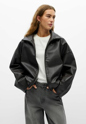 Jeune femme aux longs cheveux portant une veste en cuir noire oversize, une chemise blanche et un jean noir délavé, les mains dans les poches.