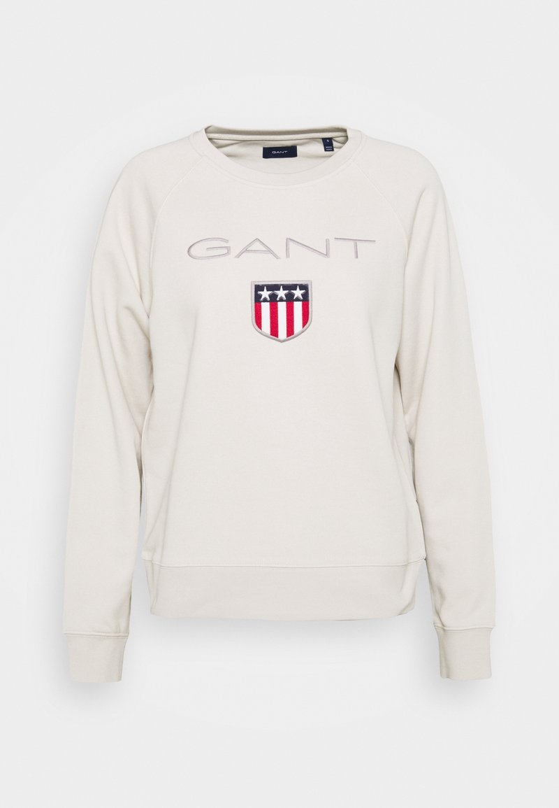 Sweat-shirt blanc cassé à col rond avec inscription "GANT" et emblème en forme de bouclier avec trois étoiles blanches au-dessus de bandes rouges et blanches sur la poitrine.
