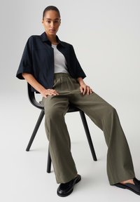 Chemise noire à boutons courte, t-shirt blanc, pantalon large vert olive et chaussures plates noires. Modèle assis sur une chaise noire.