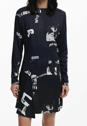 Woman wearing a black long-sleeve dress with white abstract text and graphic print, standing against a white background.