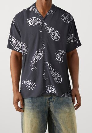 Uomo che indossa una camicia nera a maniche corte con grandi motivi paisley bianchi e jeans blu sbiaditi.