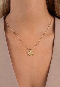 Collar de oro con un colgante redondo que presenta piedras preciosas de múltiples colores, que se lleva en el cuello de una persona, sobre una blusa de pico beige.
