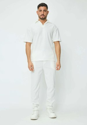 Polo shirt blanc à manches courtes avec une texture côtelée et un col évasé, associé à un pantalon blanc côtelé assorti et des baskets blanches.