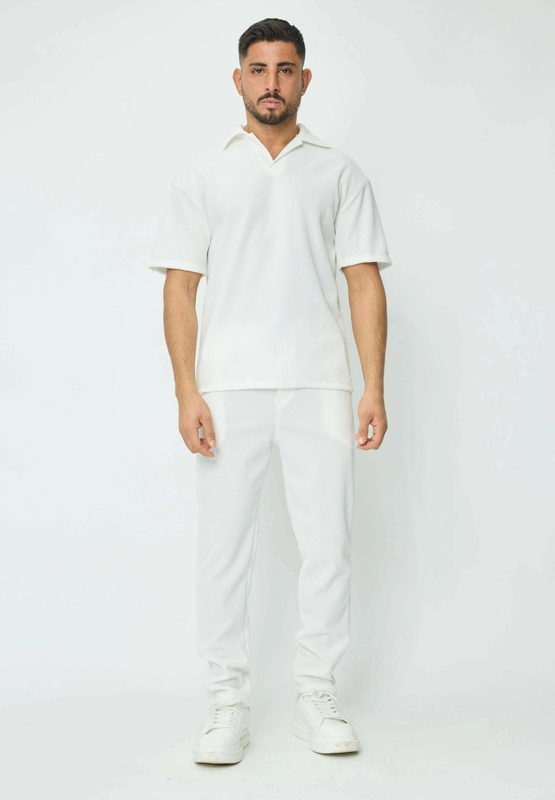 Polo shirt blanc à manches courtes avec une texture côtelée et un col évasé, associé à un pantalon blanc côtelé assorti et des baskets blanches.