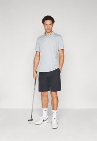 Svetlo siva polo majica s kragnom, uparena s tamnim kratkim hlačama. Model drži golf palicu, nosi bele patike i čarape s crnim detaljima.