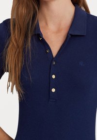 Tmavě modrá polo košile s ribbovanou texturou, třemi zlatými knoflíky a jemným vyšívaným logem na levé hrudi.