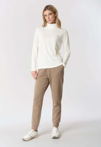 Donna con indosso un top a collo alto bianco a maniche lunghe, pantaloni da jogging beige e sneaker bianche, in piedi con una mano in tasca.