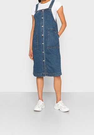 Frau trägt knielanges, blaues Jeans-Overall-Kleid mit Knöpfen, Vordertaschen und weißen Sneakers, steht vor einfachem Hintergrund.