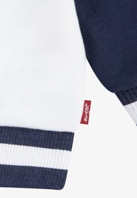 Levi's pulóver fehér testtel, sötétkék ujjakra és bordázott, Navy és fehér csíkos mandzsettával rendelkezik. Oldalán piros címke található.