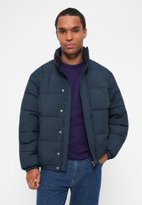 Chaqueta de plumas azul marino con diseño acolchado, cierres a presión y un cuello alto. Llevada sobre una camiseta morada, combinada con jeans azules.