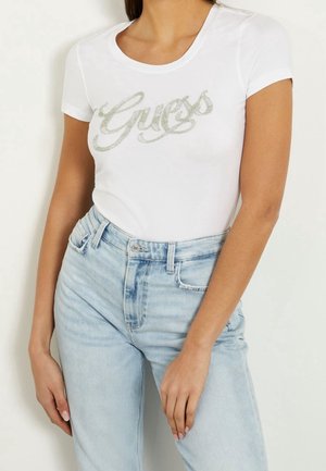 Kvinde iført en hvid t-shirt med korte ærmer med "Guess" logo i sølv glitter skrift og lyseblå højtsiddende jeans.