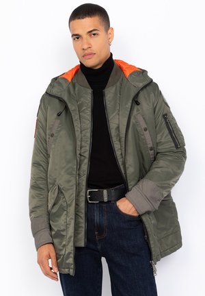 Grüne Nylonparka-Jacke mit orangefarbener Kapuzenfutter, gestepptem Muster, mehreren Reißverschlusstaschen und grauen Akzenten. Über einem schwarzen Rollkragenpullover getragen.