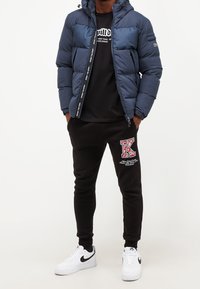 Tmavě modrá prošívaná bunda s kapucí, černé tričko s grafikou, černé tepláky s červeným vzorovaným písmenem „K“ a bílé tenisky s černými detaily.