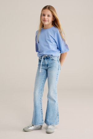 Noor tüdruk pikkade blondide juustega, kes kannab helerohelise mustriga pluusi, heledaid lõhenevaid teksaseid ja hõbedasi tossusid, seisab käed selja taga.