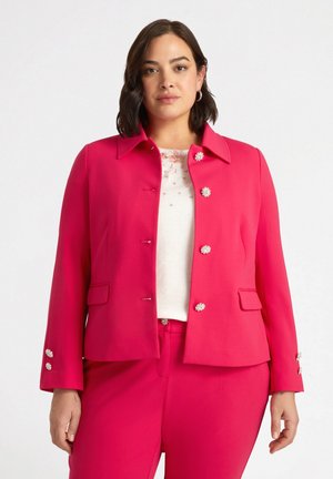 Giacca rosa brillante con colletto a pistagna, accenti di bottoni in tonalità argento e tasche laterali. Indossata sopra un top di colore chiaro con decorazioni.