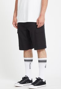 Schwarze Cargo-Shorts mit Seitentaschen, aus strapazierfähigem Material. Kombiniert mit weißen, gestreiften Volcom-Socken und schwarzen Low-Top-Sneakern.
