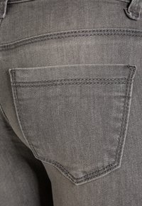 Los jeans de mezclilla gris presentan un bolsillo trasero con sutiles detalles de costura. La tela tiene una textura suave con un ligero estiramiento.