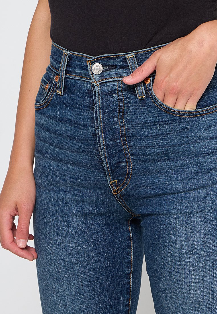 Jeans in denim blu a vita alta con un lavaggio sbiadito, cuciture arancioni e tasche frontali. Presentano una chiusura con bottone metallico e passanti per cintura.