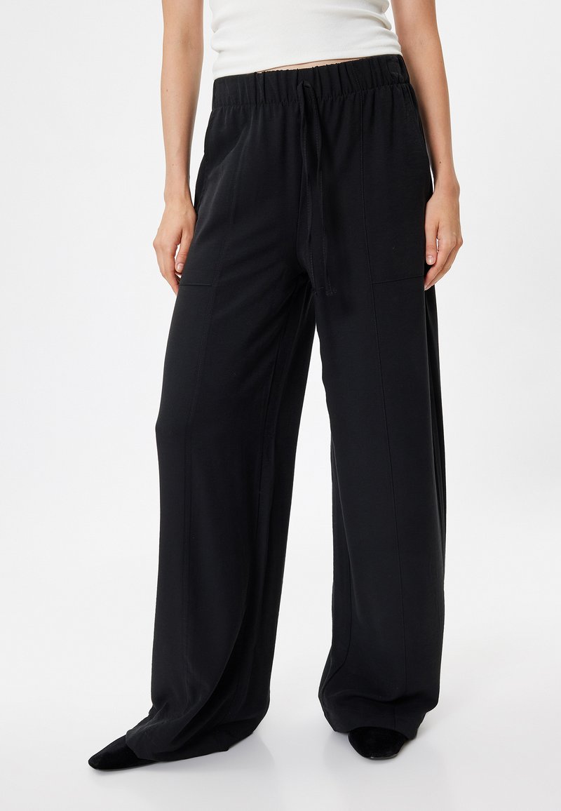 Schwarze weite Hose aus weichem Stoff, mit elastischem Bund und Kordelzug, zwei Seitentaschen und einer glatten Textur.