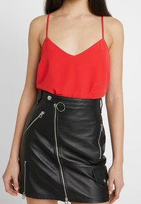Červený top s tenkými ramínky a V-výstřihem, doplněný černou, přiléhavou koženou sukní s zipy a dekorativními kovovými akcenty.